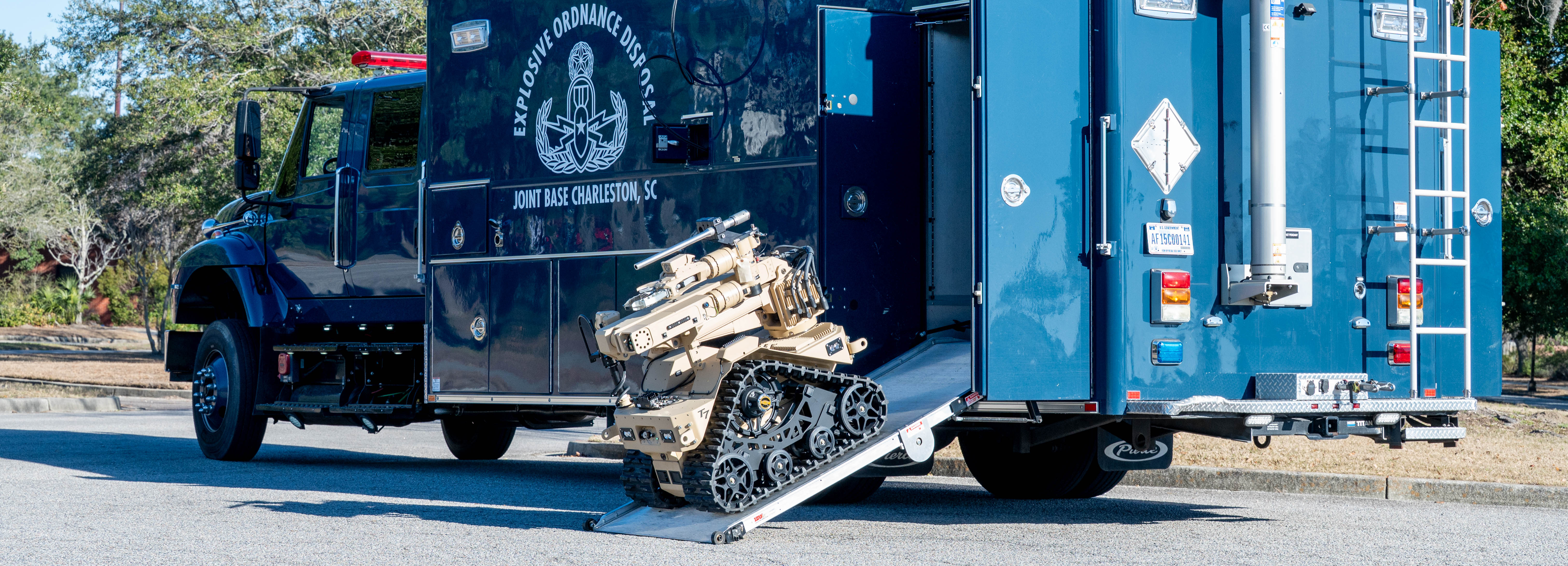 A compact, tan robot rolls down the ramp of a large blue truck with the words 'Explosive Ordnance Disposal Joint Base Charleston SC' on the side.